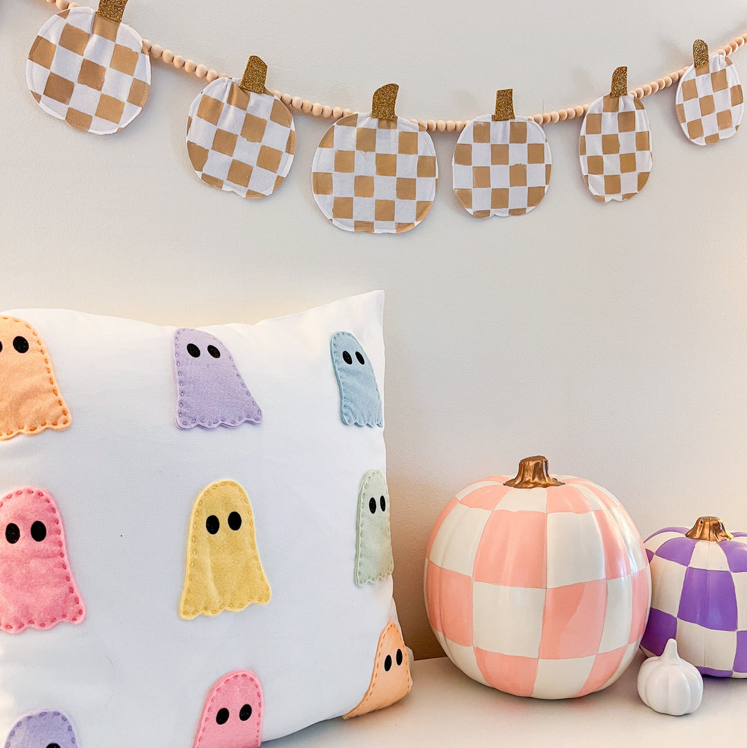 White pillow with 9 pastel colored felt ghosts, checkerboard painted white pumpkins and checkerboard pumpkin pennant hanging above it all.