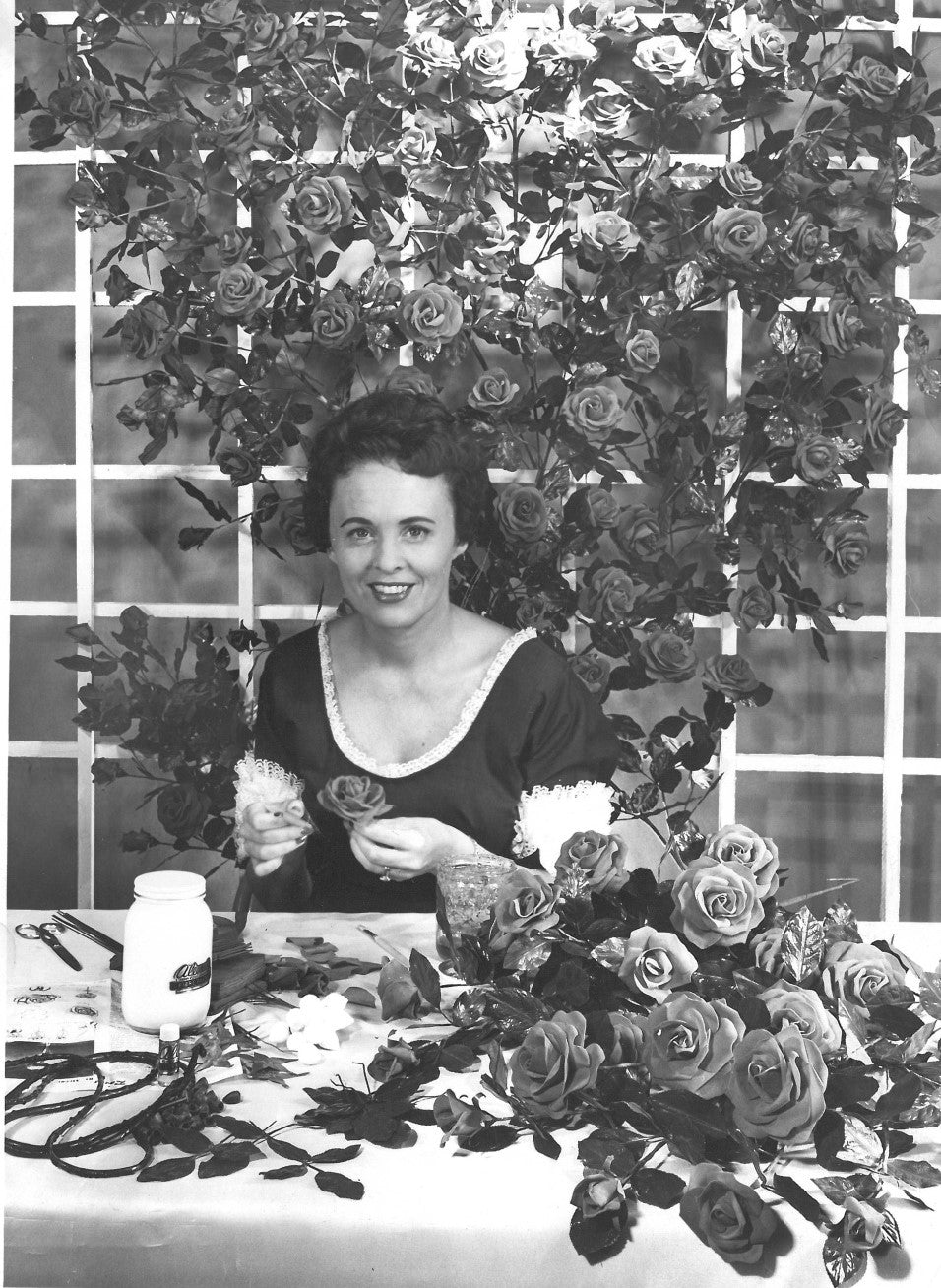 Black and white photo of a woman sitting among roses, holding a flower while crafting.