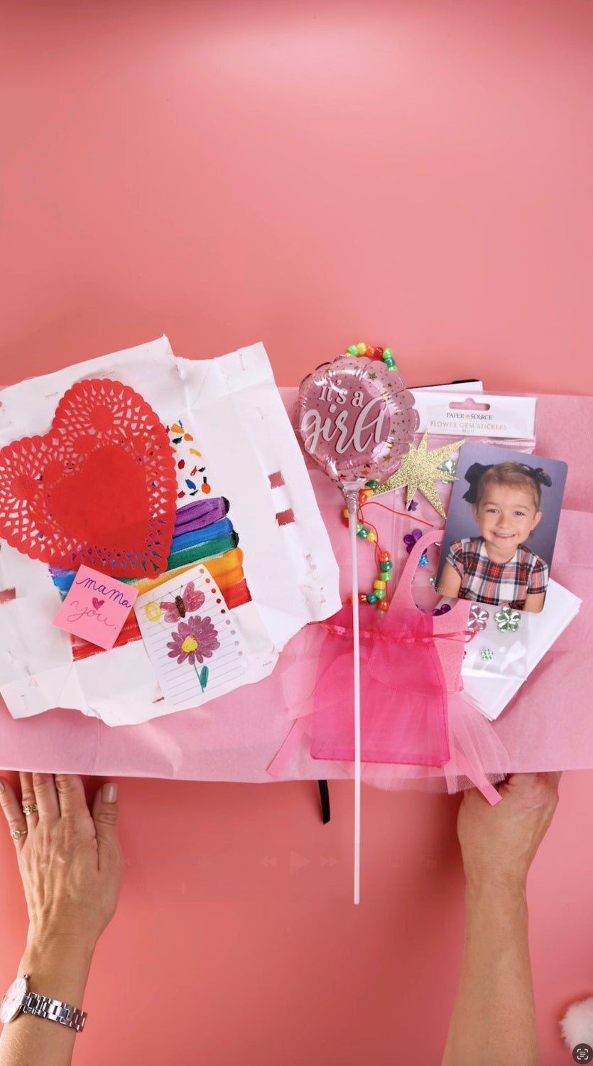 Pink background with two hands holding an open journal that has been adorned with red heart, picture of young girl, small “It’s a Girl” balloon on a stick and other mementos. 