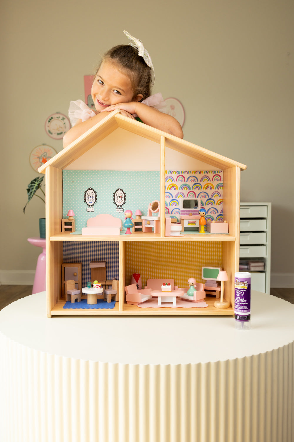 Young girl standing behind a wooden miniature dollhouse with four sections. Each section has wallpaper décor and miniature furniture depicted a bedroom and bathroom upstairs and a kitchen and family room downstairs. 
