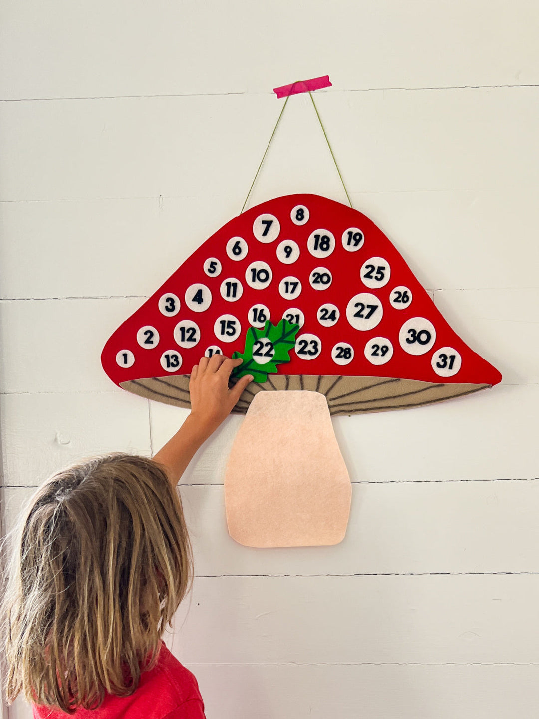 Red felt mushroom calendar with white circles & black numbers. Child reaching up to put green felt around number 22.   