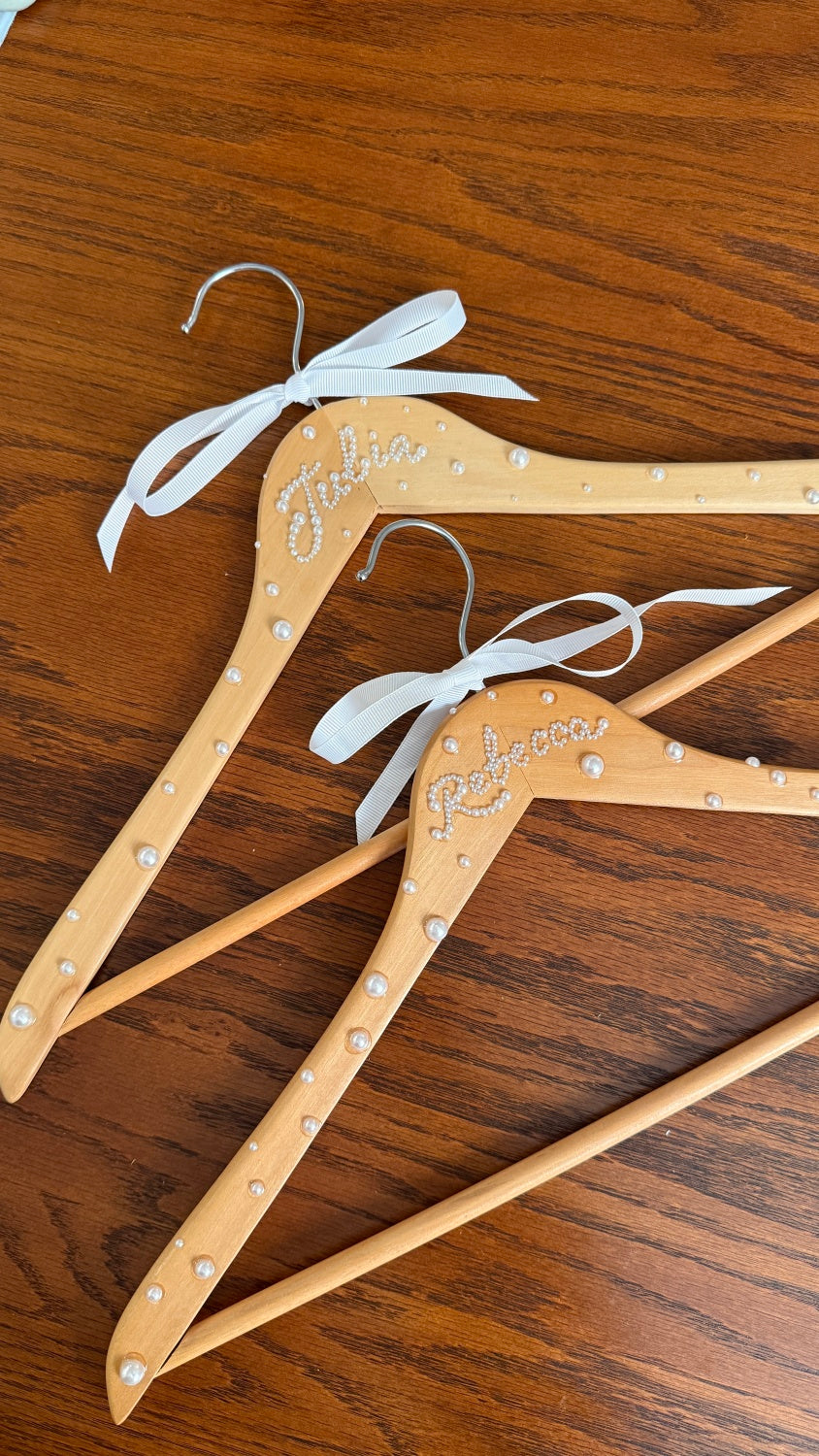 Two wooden hangers, bottle of Aleene’s Super Glue, white ribbon, wax pencil, tweezers, box of flat back pearls on a brown wooden table. 