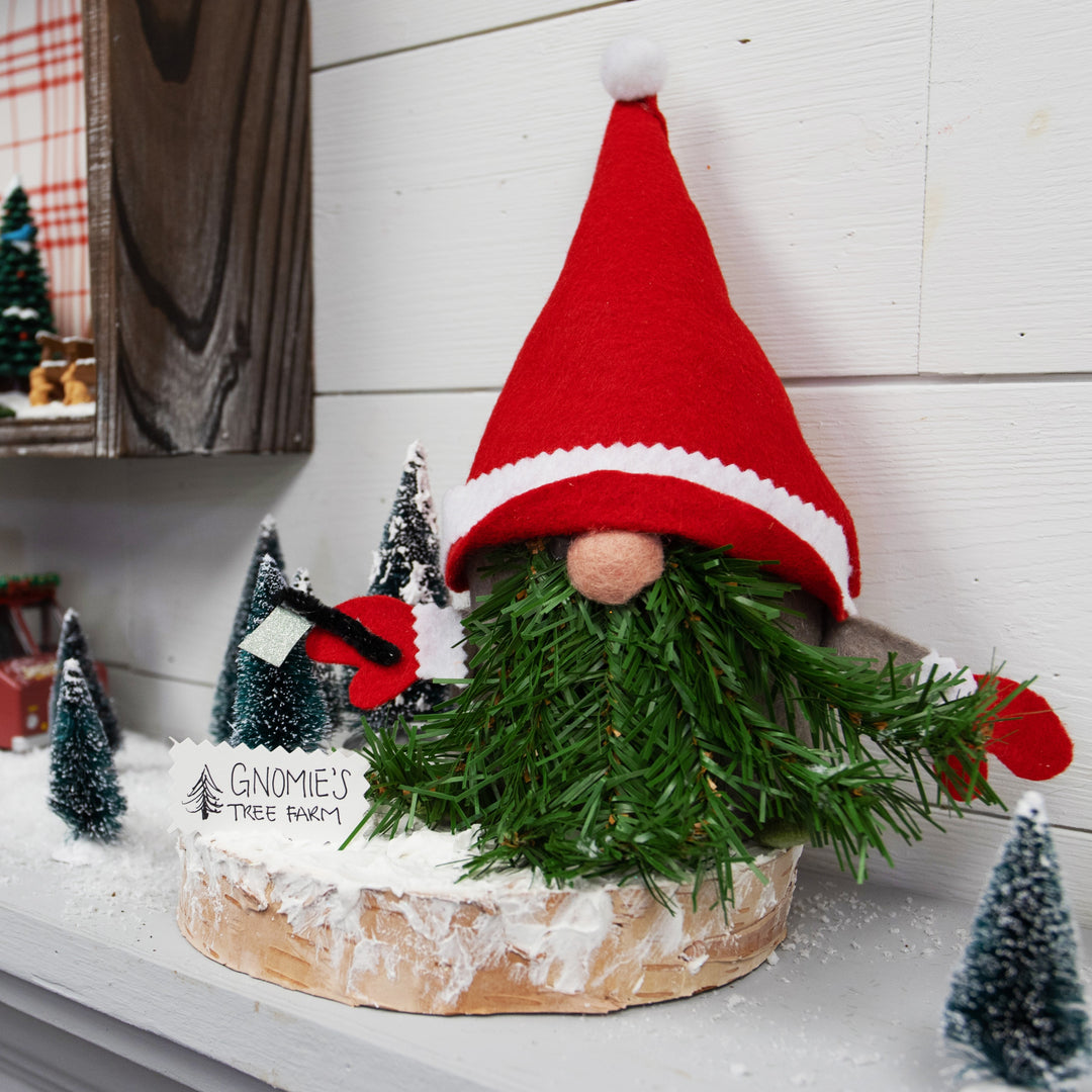 Decorative gnome with a red hat and green beard on a white surface with small Christmas trees.