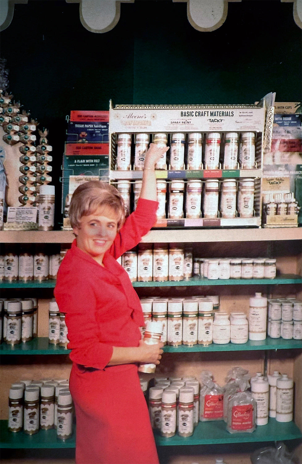 A woman in a red dress stands in front of a display of Aleene’s craft materials, reaching up to grab a bottle from the shelf while holding another in her other hand. Shelves behind her are filled with various adhesives and crafting supplies, neatly arranged. The display sign reads 'Aleene’s Basic Craft Materials' with 'Tacky' adhesive prominently featured.