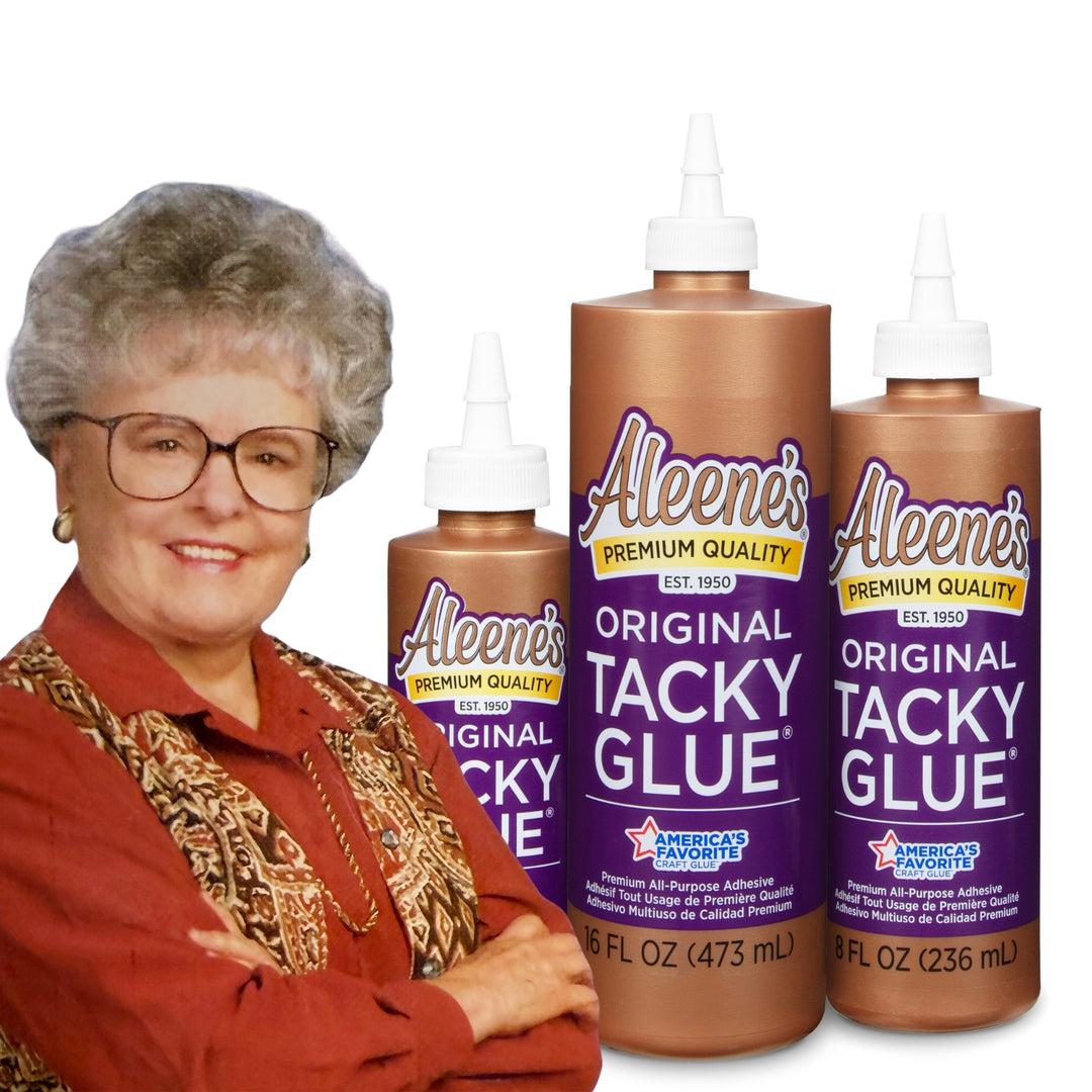 Smiling older woman with short curly hair and glasses standing with arms crossed in front of three large bottles of Aleene’s Original Tacky Glue.