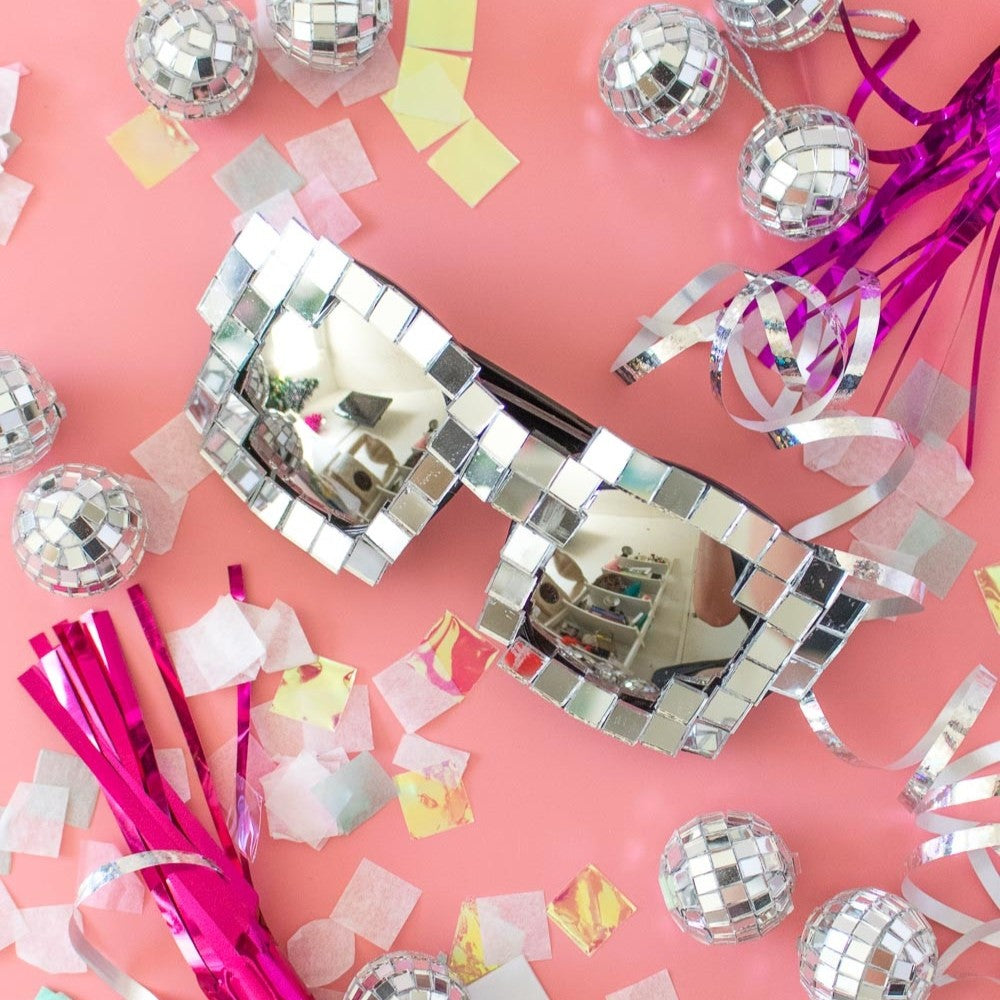 Silver disco mask surrounded by confetti and disco balls on a pink background