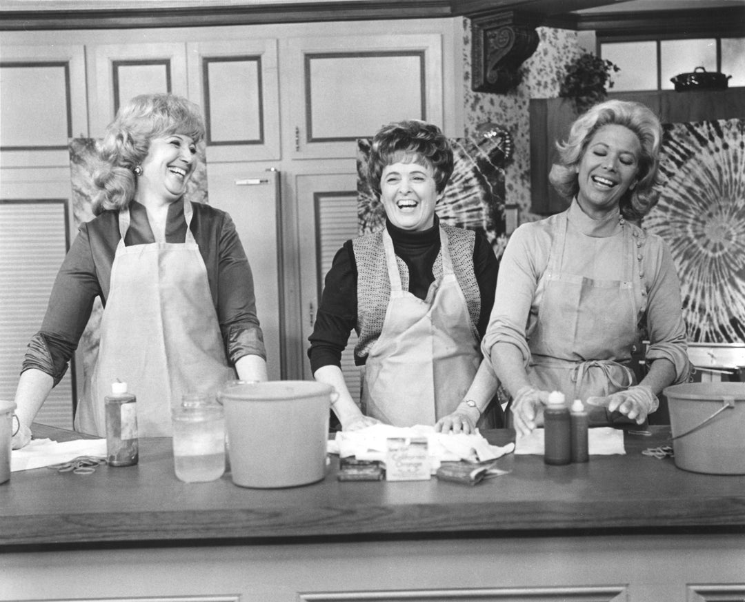 Black and white photo of three women wearing aprons, smiling while working on a craft project at a table.